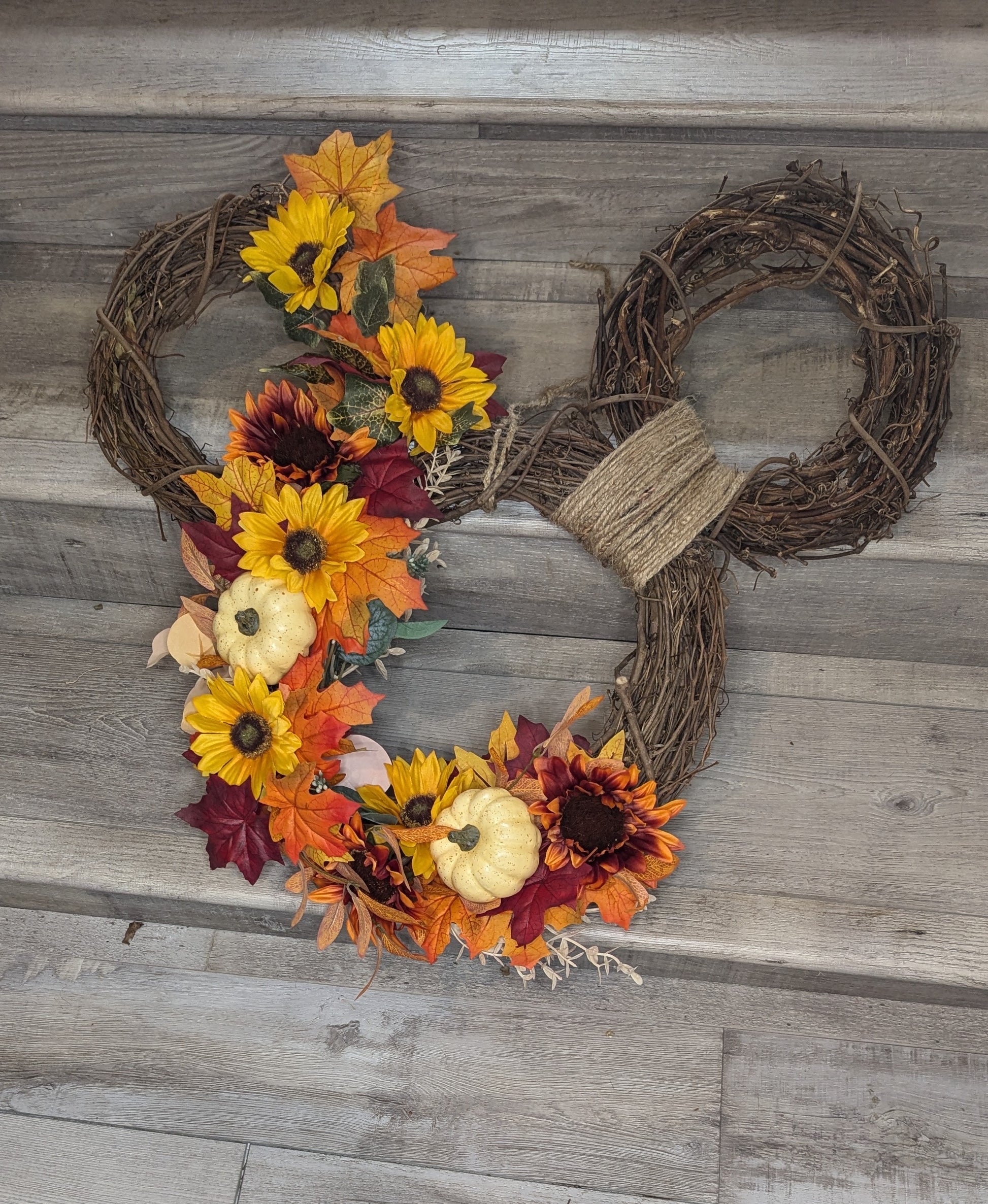 A grapevine wreath in the shape of a Mickey Mouse head outline, decorated with fall-colored leaves, sunflowers, and cream pumpkins.